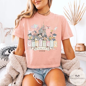 Pu&ograve; includere: T-shirt rosa chiaro con grafica colorata di personaggi Disney, un castello e la scritta "Graduate". &Egrave; incluso anche il testo "From the Tassel to the Castle 2026". La maglietta &egrave; a maniche corte.