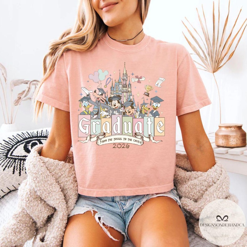 Pu&ograve; includere: T-shirt rosa chiaro con grafica colorata di personaggi Disney, un castello e la scritta "Graduate". &Egrave; incluso anche il testo "From the Tassel to the Castle 2026". La maglietta &egrave; a maniche corte.