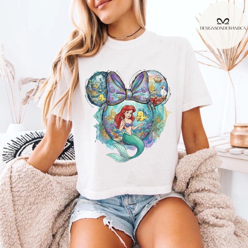 Puede incluir: Camiseta blanca con un gr&aacute;fico colorido de Ariel de La Sirenita dentro de una silueta de oreja de Mickey Mouse. El dise&ntilde;o incluye un lazo morado y escenas submarinas. La camiseta la lleva una persona.