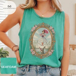 May include: A seafoam green sleeveless tank top featuring a central graphic of a person in a dress holding a pink umbrella, balancing on a tightrope above an alligator, all enclosed within an ornate gold oval frame decorated with pink roses.