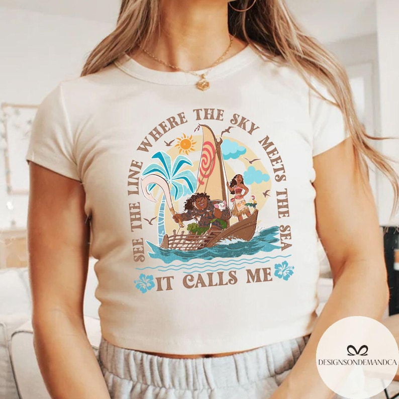 May include: Cream-colored t-shirt featuring a colorful illustration of a boat with characters, palm trees, and the sun. The text on the shirt reads "Where the sky meets the sea, see the line, it calls me."