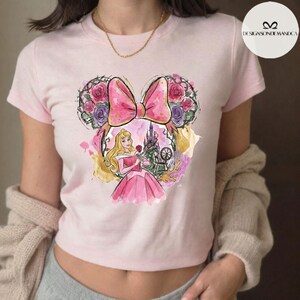Puede incluir: Camiseta rosa claro con una ilustraci&oacute;n en acuarela de la Princesa Aurora. El dise&ntilde;o incluye un lazo rosa, orejas de Mickey Mouse florales y un castillo. La camiseta es de estilo corto.
