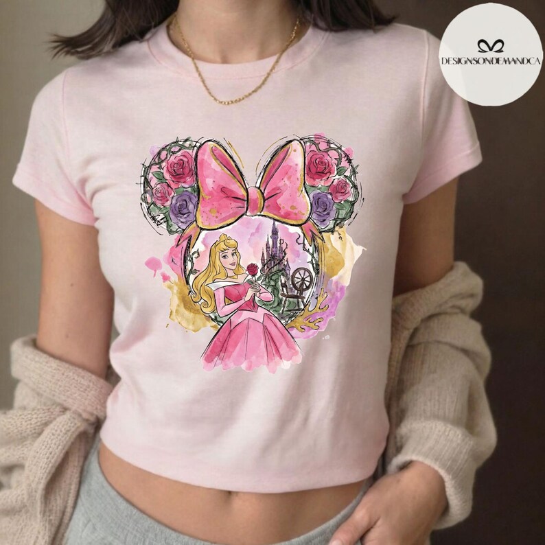 Puede incluir: Camiseta rosa claro con una ilustraci&oacute;n en acuarela de la Princesa Aurora. El dise&ntilde;o incluye un lazo rosa, orejas de Mickey Mouse florales y un castillo. La camiseta es de estilo corto.