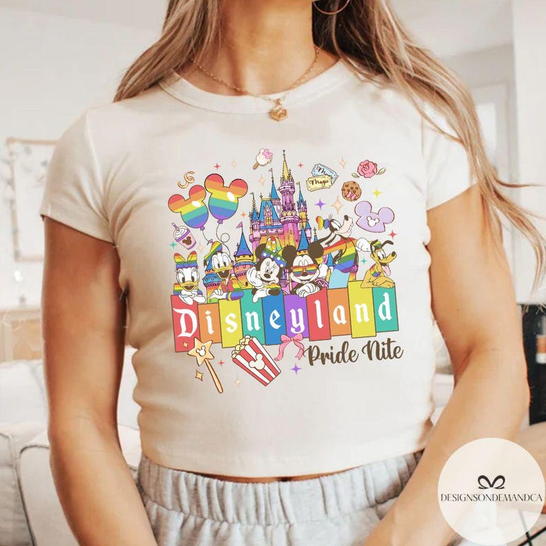 May include: Cream-coloured t-shirt with a colourful Disneyland Pride Nite design. The graphic includes Disney characters, a castle, rainbow balloons, and the text "Disneyland Pride Nite" in a vibrant font. The shirt is short-sleeved.