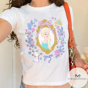 May include: White t-shirt with a colorful graphic of a princess in a frame surrounded by flowers and a snowman. The princess has long blonde hair and is wearing a blue dress. The text "Designsondemanda" is visible in the bottom right corner.