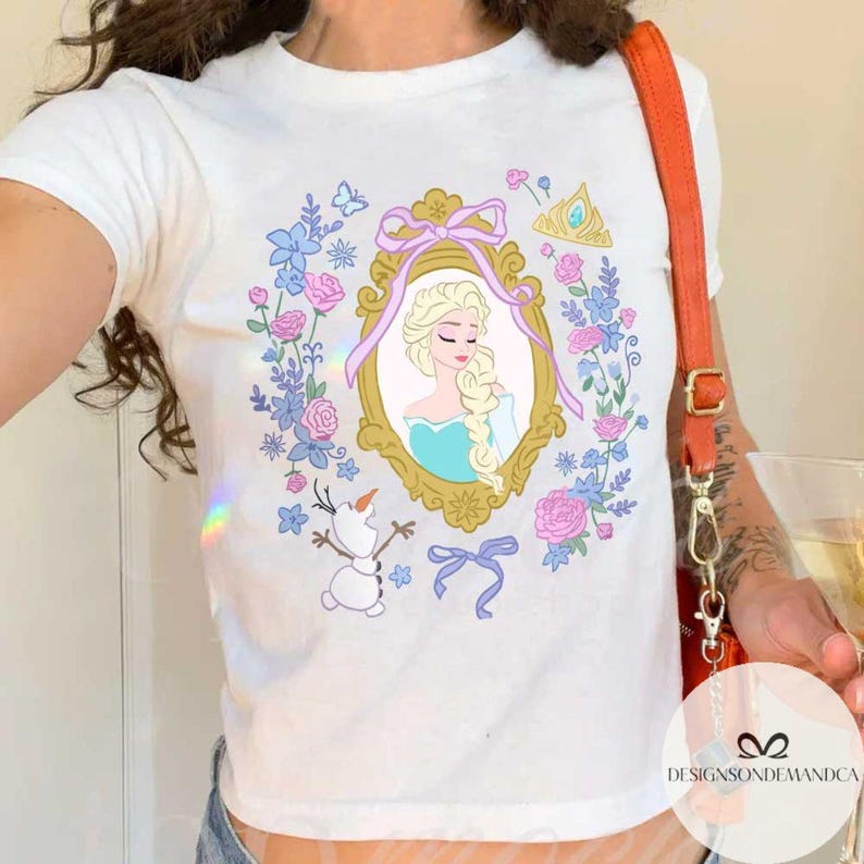 May include: White t-shirt with a colorful graphic of a princess in a frame surrounded by flowers and a snowman. The princess has long blonde hair and is wearing a blue dress. The text "Designsondemanda" is visible in the bottom right corner.
