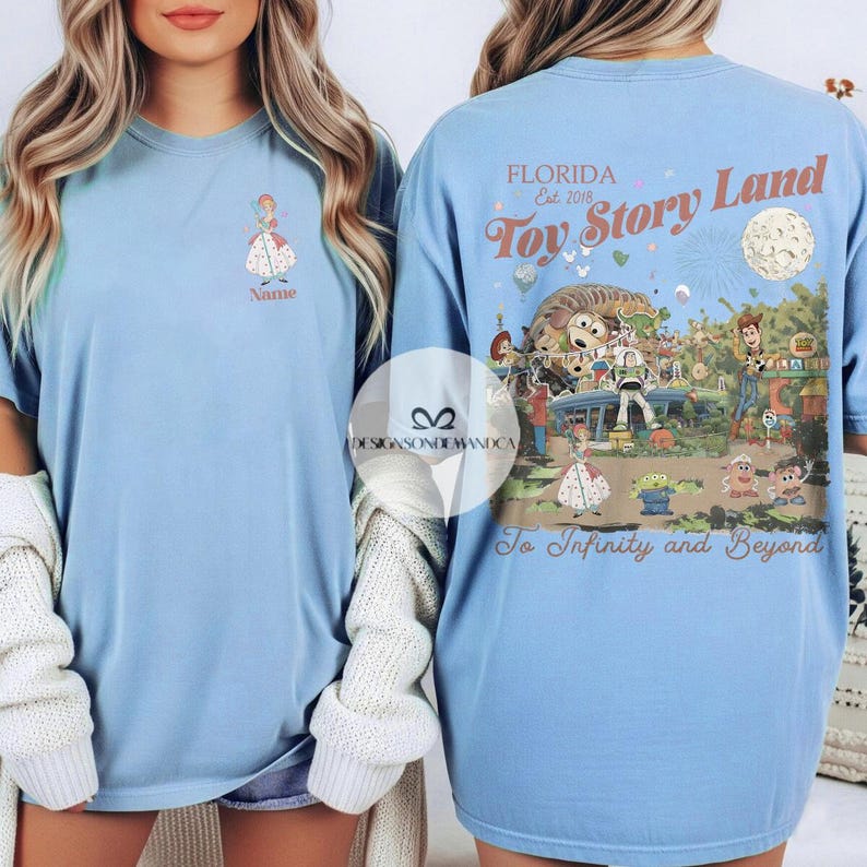 Puede incluir: Camiseta azul claro con un gr&aacute;fico de dibujos animados en la parte delantera y trasera. La parte delantera tiene un gr&aacute;fico de princesa con la palabra "Name" debajo. La parte trasera tiene un dise&ntilde;o de "Toy Story Land" con el texto "To Infinity and Beyond".