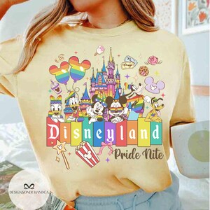 May include: A pale yellow t-shirt with a Disneyland Pride Nite design. The graphic features Mickey and Minnie Mouse, Donald Duck, and Pluto, alongside a castle, balloons, and the Disneyland logo in rainbow colours.