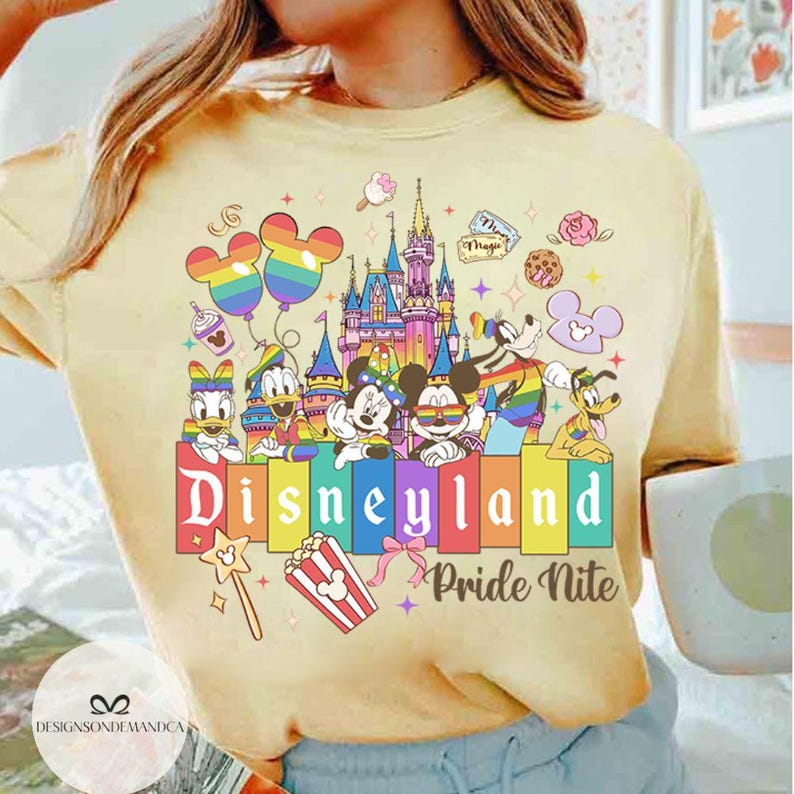 May include: A pale yellow t-shirt with a Disneyland Pride Nite design. The graphic features Mickey and Minnie Mouse, Donald Duck, and Pluto, alongside a castle, balloons, and the Disneyland logo in rainbow colours.