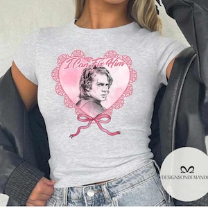 May include: A gray t-shirt with a pink heart design featuring a drawing of a man with the text "I Can Fix Him" inside the heart.