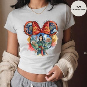 Puede incluir: Camiseta gris claro con un gr&aacute;fico colorido de Mulan en un dise&ntilde;o de oreja de Mickey Mouse. El gr&aacute;fico incluye un lazo rojo, patrones de inspiraci&oacute;n china y una ilustraci&oacute;n de acuarela de Mulan con una espada, la Gran Muralla y un palacio.