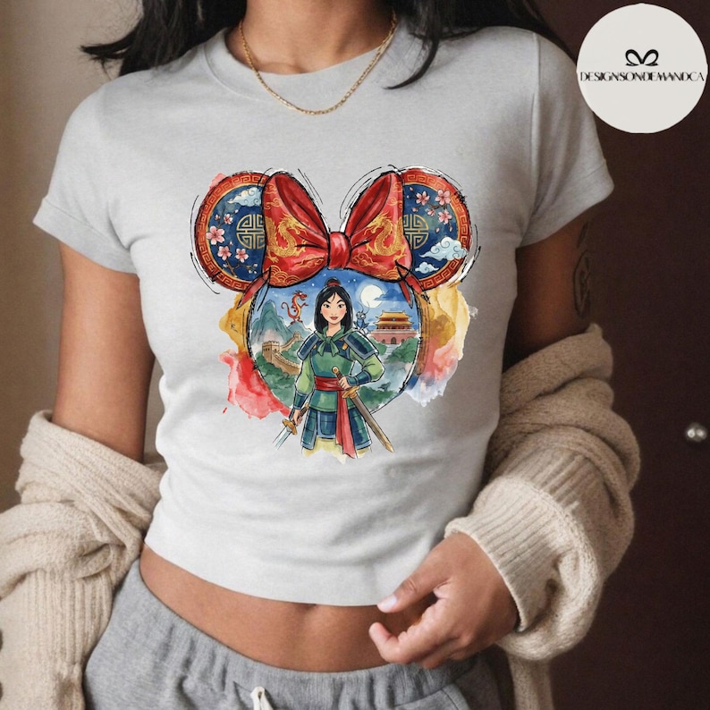 Puede incluir: Camiseta gris claro con un gr&aacute;fico colorido de Mulan en un dise&ntilde;o de oreja de Mickey Mouse. El gr&aacute;fico incluye un lazo rojo, patrones de inspiraci&oacute;n china y una ilustraci&oacute;n de acuarela de Mulan con una espada, la Gran Muralla y un palacio.