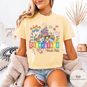 May include: Pale yellow t-shirt with a vibrant Disneyland graphic featuring Disney characters. The text "Disneyland Pride Nite" is displayed below the graphic. The shirt has short sleeves and a crew neck. The design includes rainbow-colored elements.