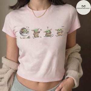 May include: A light pink t-shirt featuring a cartoon of a character in various poses. The character is green with large ears and is wearing a tan robe. The word "cool" is written on the shirt.