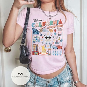 以下が含まれることがあります： ディズニー・カリフォルニア・アドベンチャーのカラフルなグラフィックが特徴のピンクのTシャツ。デザインにはミッキーマウス、観覧車、ジェットコースター、その他のパークアトラクションが含まれています。Tシャツは半袖で、クルーネックです。