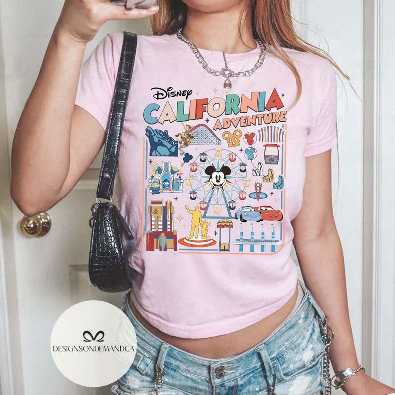 以下が含まれることがあります： ディズニー・カリフォルニア・アドベンチャーのカラフルなグラフィックが特徴のピンクのTシャツ。デザインにはミッキーマウス、観覧車、ジェットコースター、その他のパークアトラクションが含まれています。Tシャツは半袖で、クルーネックです。