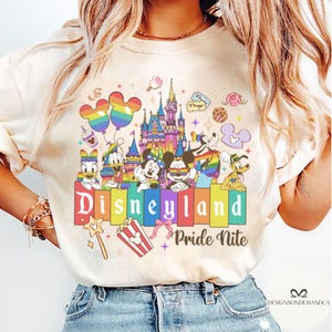May include: A cream-coloured t-shirt with a Disneyland Pride Nite design. The graphic features Mickey and Minnie Mouse, Donald Duck, Goofy, and Pluto, a rainbow castle, balloons, and the Disneyland logo in a rainbow colour scheme.
