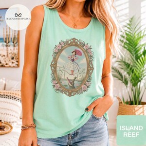 May include: A light mint green sleeveless tank top featuring a central graphic of a person in a dress holding a pink umbrella while balancing on a tightrope above an alligator, all enclosed within an ornate, rose-adorned oval frame.