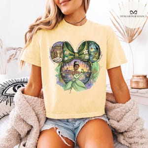 Puede incluir: Camiseta amarilla clara con un dise&ntilde;o de la Princesa Tiana en forma de oreja de Mickey Mouse. El dise&ntilde;o incluye un lazo verde, un paisaje urbano e im&aacute;genes de una rana y flores.