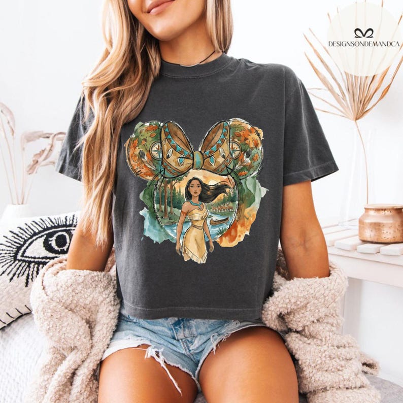 Puede incluir: Camiseta gris oscuro con un gr&aacute;fico colorido de una princesa nativa americana dentro de un dise&ntilde;o de oreja de Mickey Mouse. El dise&ntilde;o incluye un lazo, &aacute;rboles y un r&iacute;o. La camiseta es de un estilo c&oacute;modo e informal.