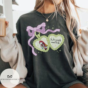 Puede incluir: Sudadera gris oscuro con un dise&ntilde;o de medall&oacute;n en forma de coraz&oacute;n, con un personaje de dibujos animados y el texto "We're simply meant to be". El medall&oacute;n est&aacute; adornado con un lazo y una cinta morados. La sudadera tiene cuello redondo.