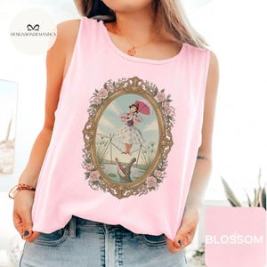 May include: A light pink sleeveless tank top featuring a central graphic print of a person balancing on a tightrope with an umbrella above an alligator, all enclosed within an ornate gold-colored oval frame decorated with pink roses.