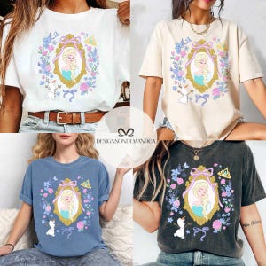 May include: Four different color t-shirts featuring a cartoon image of Elsa from Frozen. The image shows Elsa in a blue dress with a crown and a floral design around her. The t-shirts are white, beige, blue, and black.