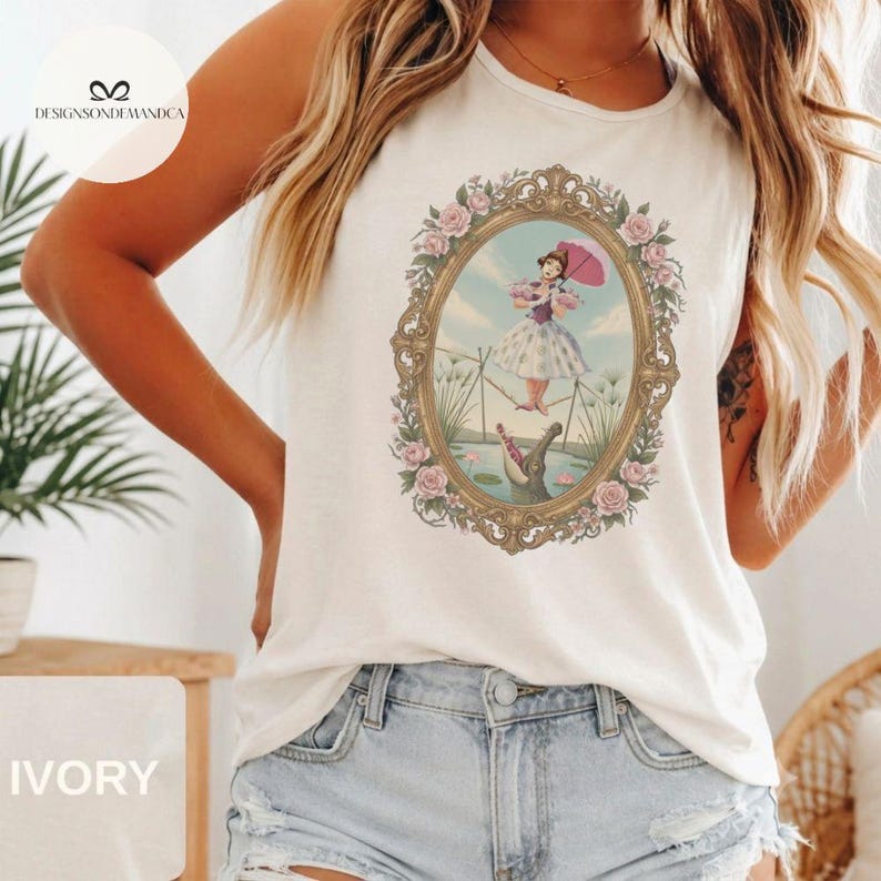 May include: An ivory sleeveless tank top featuring a central graphic of a person in a dress holding a pink umbrella while tightrope walking above an alligator. The scene is enclosed in an ornate gold oval frame decorated with pink roses.