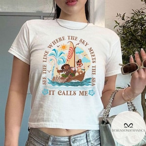 May include: White t-shirt featuring a colorful illustration of a boat with characters and the text "SEE THE LINE WHERE THE SKY MEETS THE SEA IT CALLS ME". The design includes a sun, palm trees, and ocean waves.