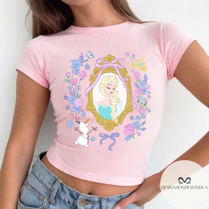 May include: Pink cropped t-shirt with a floral design featuring a cartoon image of Elsa from Frozen in a gold frame. The design also includes a cartoon image of Olaf and a crown.
