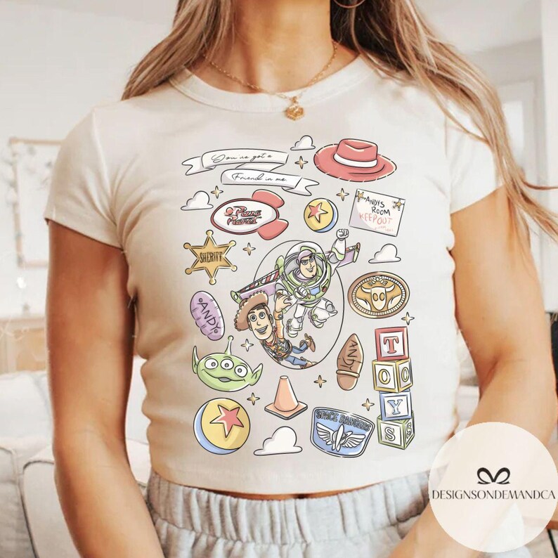 May include: Cream-colored crop top featuring a colorful graphic design of characters and elements from the animated film Toy Story. The design includes Woody, Buzz Lightyear, and other iconic imagery. The text "Gonna get a friend in me" is also visible.