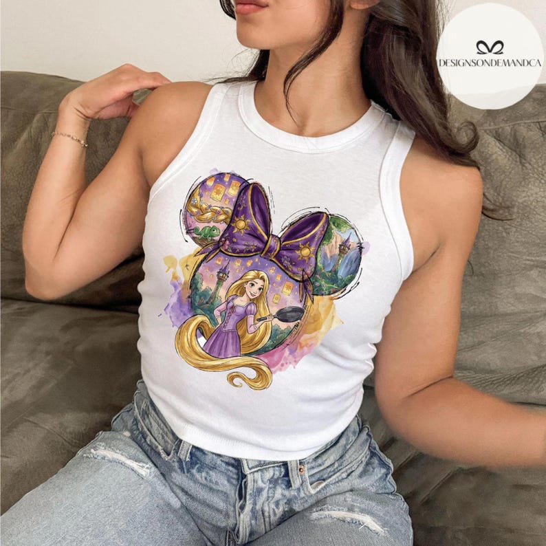 Puede incluir: Camiseta sin mangas blanca con un dise&ntilde;o de lazo morado y una ilustraci&oacute;n de dibujos animados de una princesa con pelo largo y rubio sosteniendo una sart&eacute;n. El dise&ntilde;o incluye un castillo y linternas flotantes.