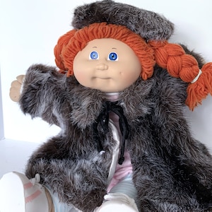 May include: A doll with bright orange yarn hair styled in two braids and a matching hat. The doll features blue eyes, a round face, and wears a dark brown faux fur coat with a black tie closure over a light-colored outfit and white shoes.