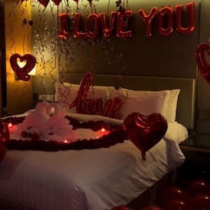 Könnte beinhalten: Ein romantisches Schlafzimmer mit einem weißen Bett, roten herzförmigen Ballons und einem Banner mit der Aufschrift "I LOVE YOU". Das Bett ist mit roten Rosenblättern und weißen Schwänen dekoriert.