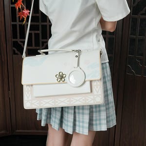 Pu&ograve; includere: Una borsa a tracolla bianca e rosa con un motivo floreale e un ciondolo circolare trasparente. La borsa ha una tracolla lunga e un manico superiore.