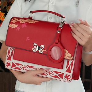 Pu&ograve; includere: Una borsa a mano in pelle rossa con un design geometrico bianco e rosso. La borsa ha un ciondolo a forma di farfalla e un ciondolo circolare con una pietra rossa. La borsa ha un manico superiore e una tracolla rimovibile.