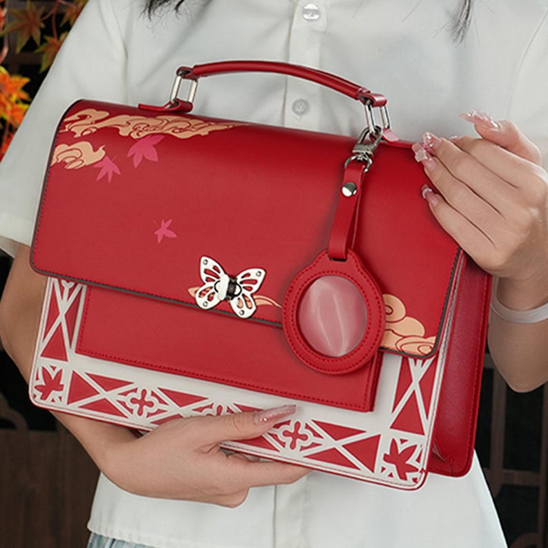 Pu&ograve; includere: Una borsa a mano in pelle rossa con un design geometrico bianco e rosso. La borsa ha un ciondolo a forma di farfalla e un ciondolo circolare con una pietra rossa. La borsa ha un manico superiore e una tracolla rimovibile.