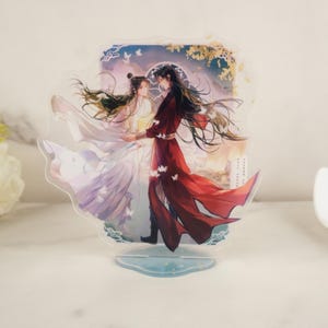 May include: A clear acrylic stand with a romantic illustration of two figures in flowing robes. The figures are embracing, set against a sunset and butterflies. The stand has a light blue base.