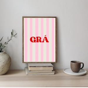 Könnte beinhalten: Gerahmter Kunstdruck mit rosa-weiß gestreiftem Hintergrund und dem roten Wort "GRÁ". Das Kunstwerk wird auf einer Holzoberfläche mit einem Stapel Bücher und einer Kaffeetasse präsentiert. Der Rahmen ist aus dunklem Holz.