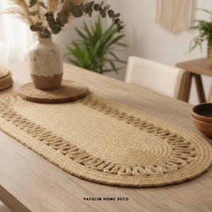 Puede incluir: Camino de mesa ovalado de yute tejido en color beige natural. El camino de mesa presenta un borde decorativo y está colocado sobre una mesa de madera. También se ven un posavasos a juego y un jarrón con plantas secas.