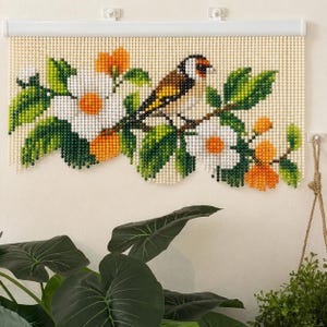 May include: A beaded wall hanging featuring a goldfinch perched on a branch with white flowers, green leaves, and orange accents. The design is composed of small, round beads arranged in a grid pattern, suspended from a white horizontal rod.