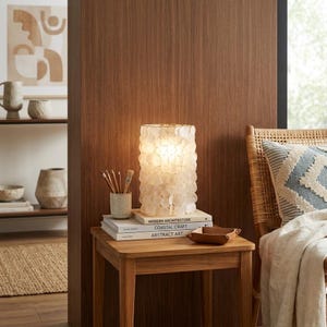 May include: A cylindrical table lamp constructed from overlapping translucent white shell discs, emitting a warm glow. It sits on a stack of three books titled Modern Architecture, Coastal Craft, and Abstract Art, next to a pencil holder.