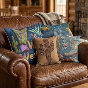 Pode incluir: Uma coleção de almofadas decorativas com tema Western. As almofadas apresentam desenhos bordados de cactos, botas de cowboy e padrões florais em tons de azul, castanho e verde. As almofadas estão dispostas num sofá de couro castanho.