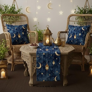 Op de afbeelding: Een decoratieve buitenscène met twee rieten stoelen en een houten tafel. De tafel is versierd met een marineblauwe tafelloper en twee bijpassende kussens, allemaal met een hemels ontwerp van manen, sterren en lantaarns. Een gouden lantaarn en theepot dragen bij aan de sfeer.