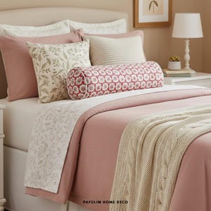 Op de afbeelding: Een bed met een blush roze dekbed en een crèmekleurige kabelgebreide deken. Decoratieve kussens zijn onder meer een bloemenprint, effen roze en een crèmekleurig gebreid kussen. Er is ook een cilindrisch kussen met een rood bloemmotief. De tekst "PAFULIM HOME DECO" is zichtbaar.