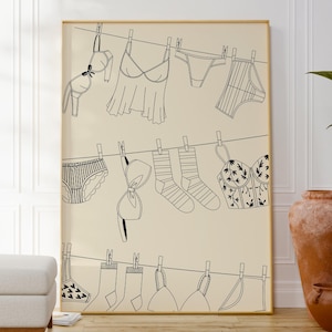 Puede incluir: Una impresión enmarcada que presenta un dibujo lineal de lencería y calcetines colgados en un tendedero. La obra de arte incluye sujetadores, ropa interior y calcetines, todos representados en líneas negras sobre un fondo crema. La impresión se exhibe en una habitación con tonos neutros.