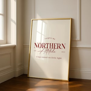 Framed typography print with the text “Forgive my northern attitude, I was raised on little light” leaning against a wall in a sunlit room with light wood floors.