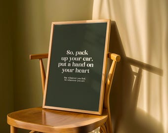 May include: A framed art print with a dark green background and the text "So, pack up your car, put a hand on your heart." The print is displayed on a wooden chair. The frame is a light brown color.
