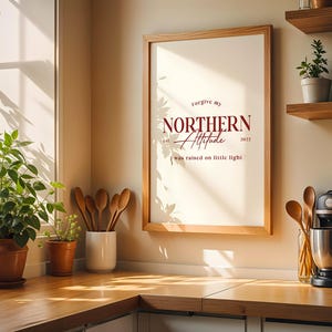 Framed typography print placed on a kitchen counter with plants, shelves, and sunlight coming through a window.