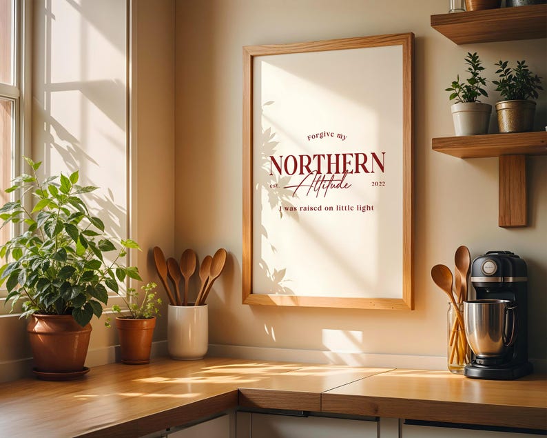 Framed typography print placed on a kitchen counter with plants, shelves, and sunlight coming through a window.